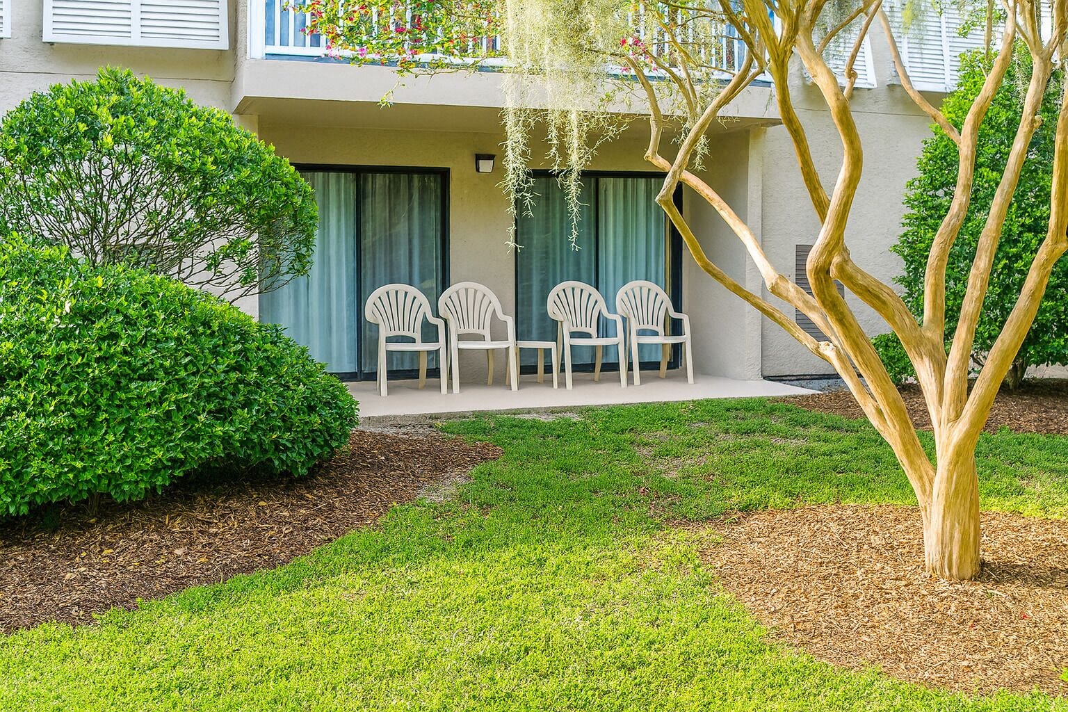 Un'area esterna curata con prato verde, un cespuglio ben rifinito e un albero. Tre sedie bianche sono disposte di fronte a un edificio con un balcone.