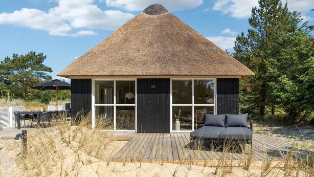 Una casa con techo de paja, ventanas amplias, una terraza de madera y un área de descanso.