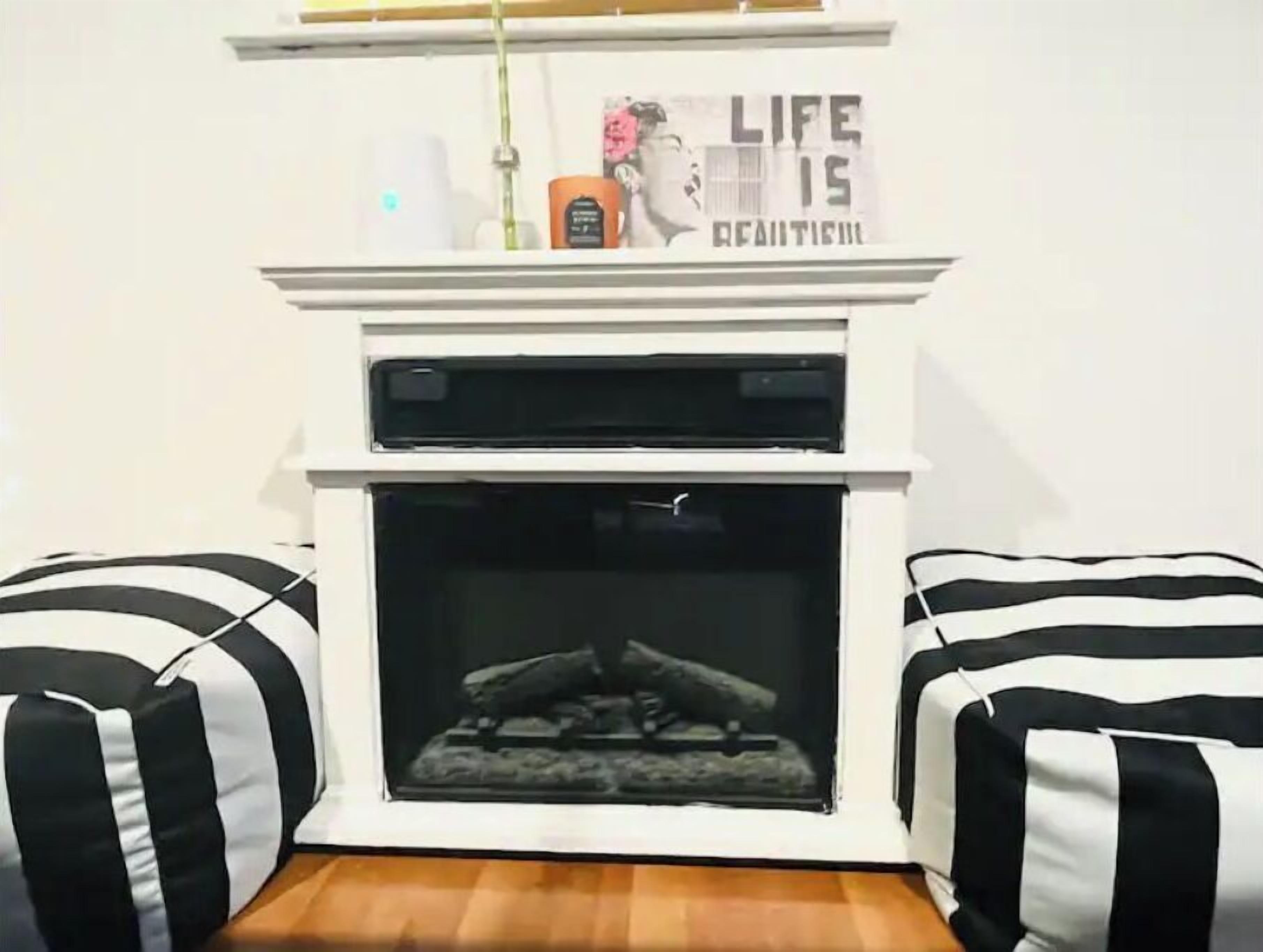 Un salon moderne avec une cheminée blanche, des coussins rayés noirs et blancs et une affiche motivante accrochée au-dessus de la cheminée.