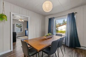 En matplats med ett träbord och stolar, en hängande växt och utsikt över utomhusmiljön genom ett fönster med gardiner.
