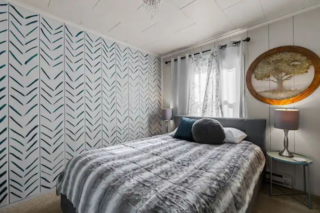 Une chambre à coucher comprenant un lit, une table de chevet, une lampe, une fenêtre avec des rideaux et un mur orné d’un motif en chevrons.
