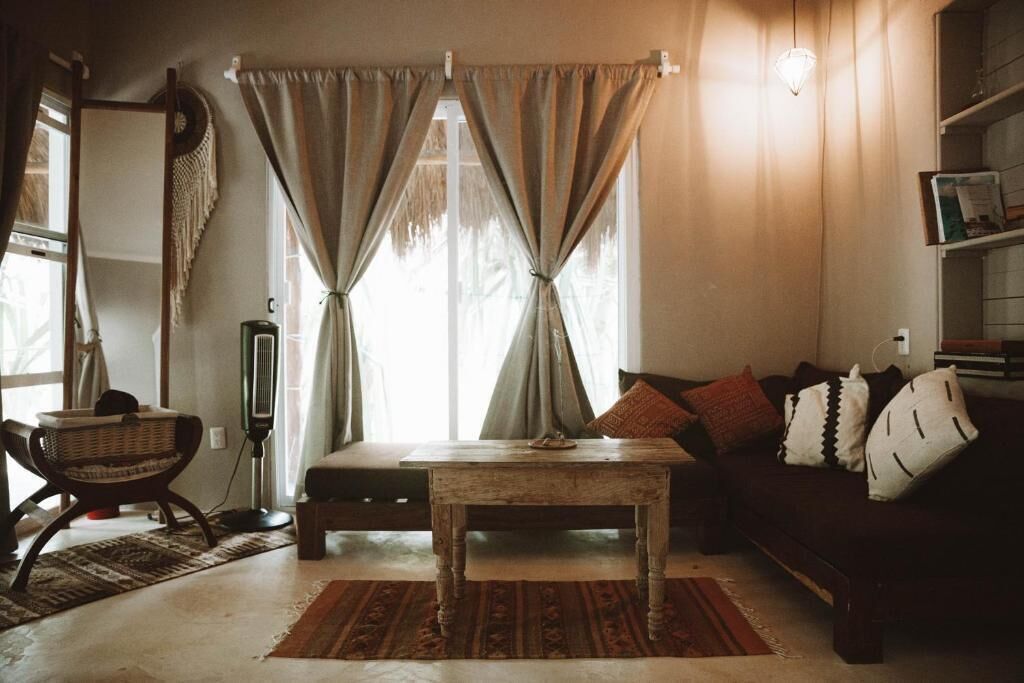 A cozy living room with a sofa, a coffee table, and a chair. There is a window with curtains, a lamp, and a bookshelf.
