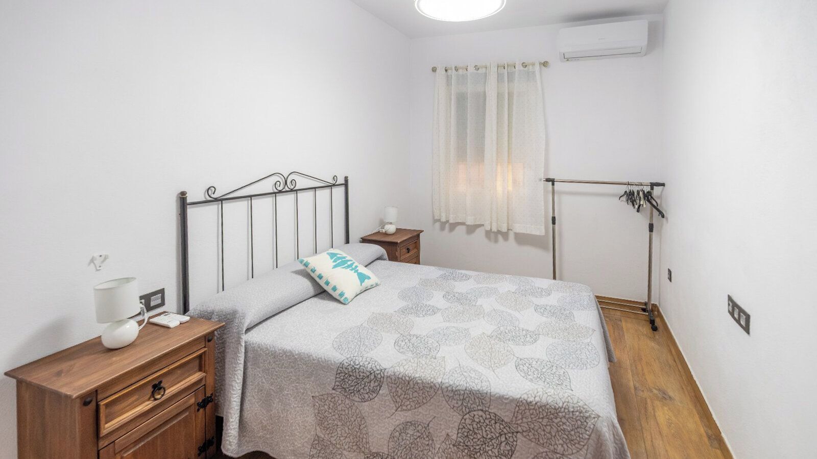 A bedroom with a bed, a nightstand, a dresser, a window with curtains, and an air conditioning unit.
