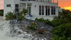 A two-story white house with a balcony, surrounded by greenery and a sandy area. - Ocean Front facing the Tropic of Cancer Beach !! (Little Exuma)