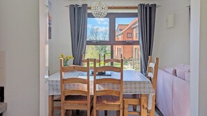 A dining area with a table and chairs, a chandelier, and a view of a brick house through large windows. - Cosy cottage with designated parking. (Whitby)