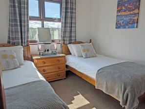 Quarto com duas camas de solteiro, cômoda de madeira, mesa de cabeceira com luminárias, janela com cortinas e um quadro na parede.