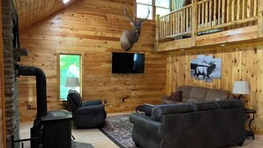 A log cabin interior with a fireplace, a television mounted on the wall, a sofa, and a deer head mounted on the wall.