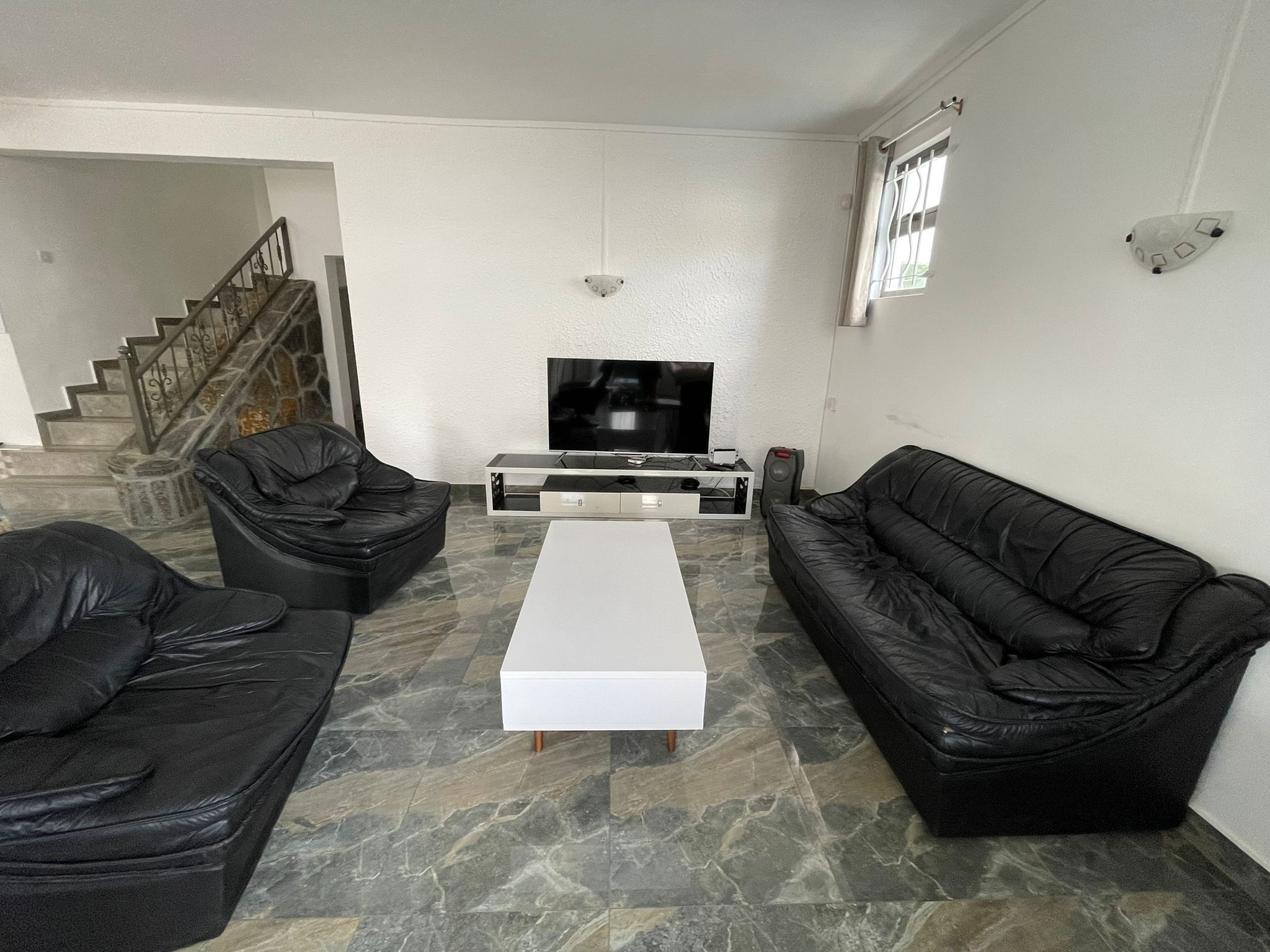 A living room with a TV, two black leather sofas, a white coffee table, and a staircase in the background.