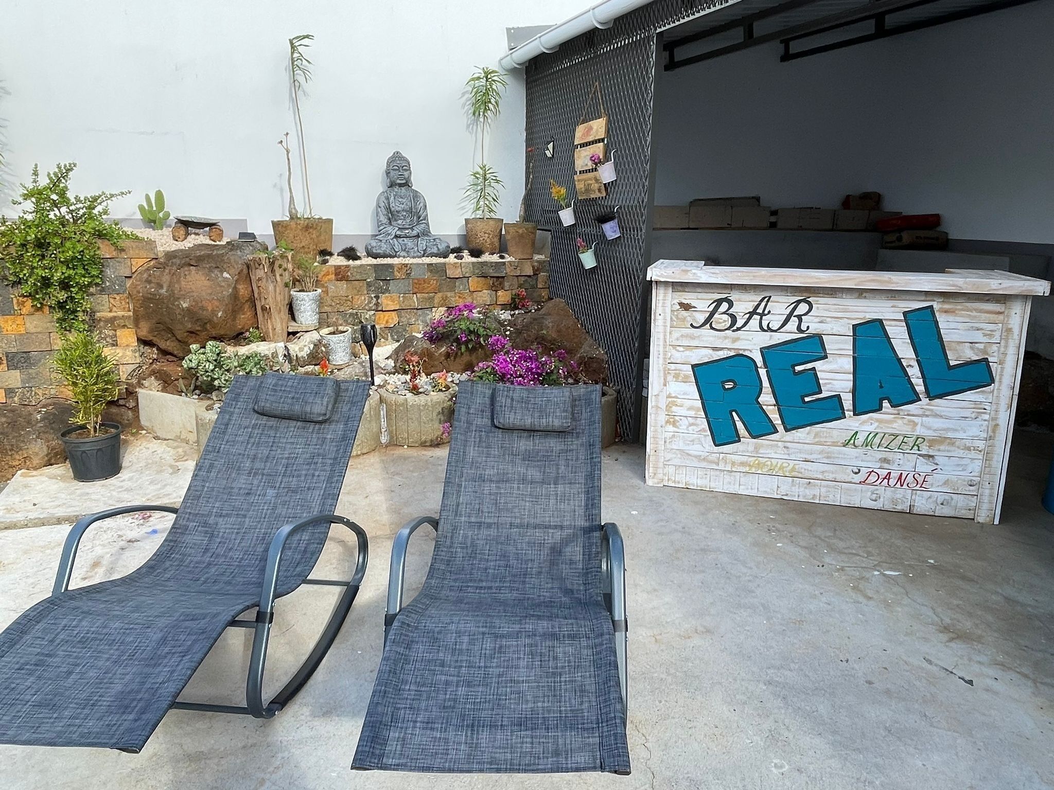 A patio with two lounge chairs, a Buddha statue, potted plants, and a wooden sign that reads "Bar REAL AMIZER DANSE".