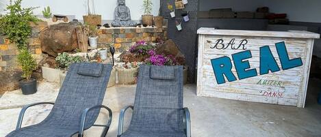 Une terrasse avec deux chaises longues, une statue de Bouddha, des plantes en pot et une enseigne en bois sur laquelle on peut lire : « Bar REAL AMIZER DANSE ».