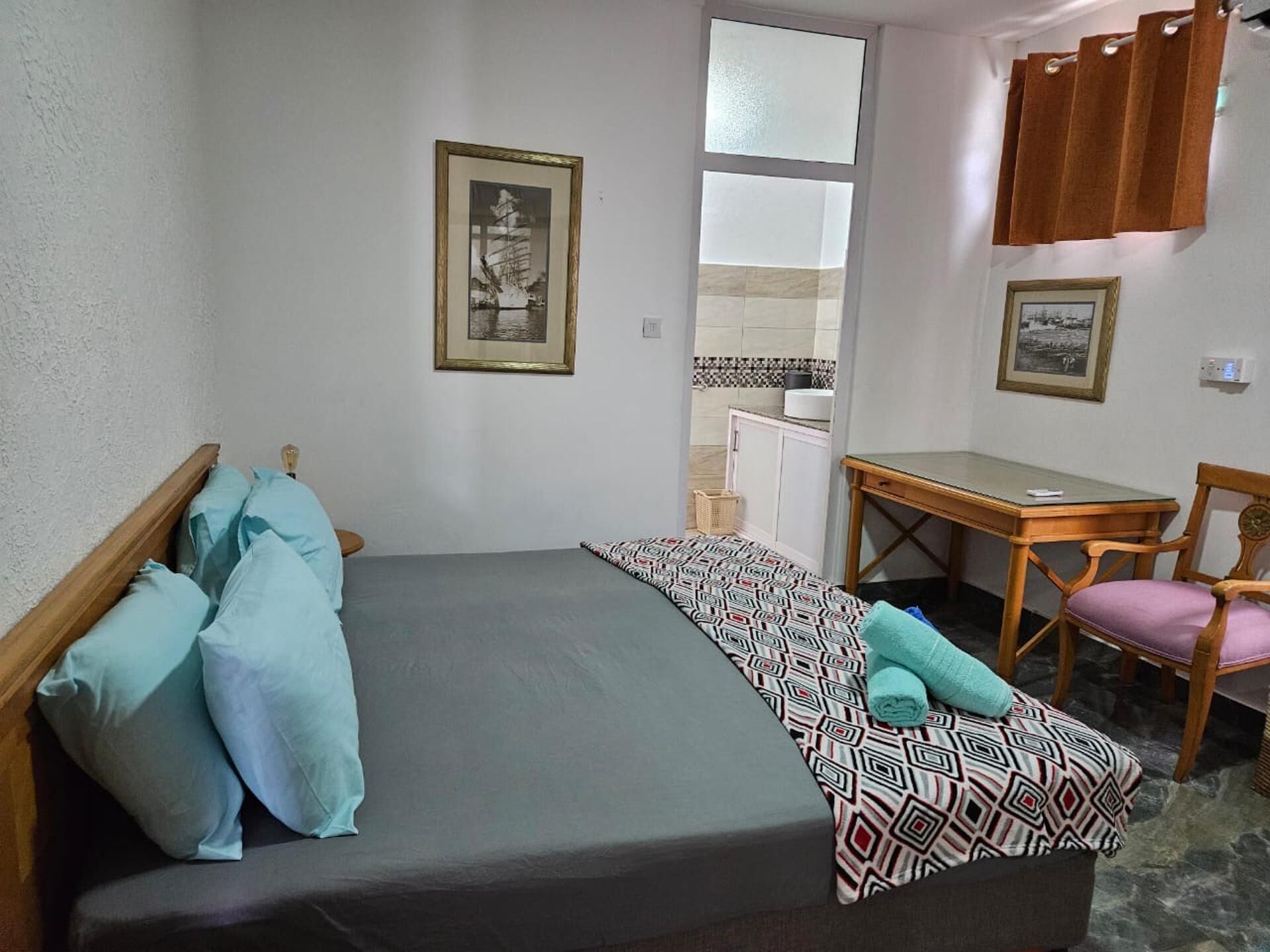Une petite pièce, bien rangée, avec un lit, un bureau et une chaise. Deux tableaux encadrés ornent le mur.
