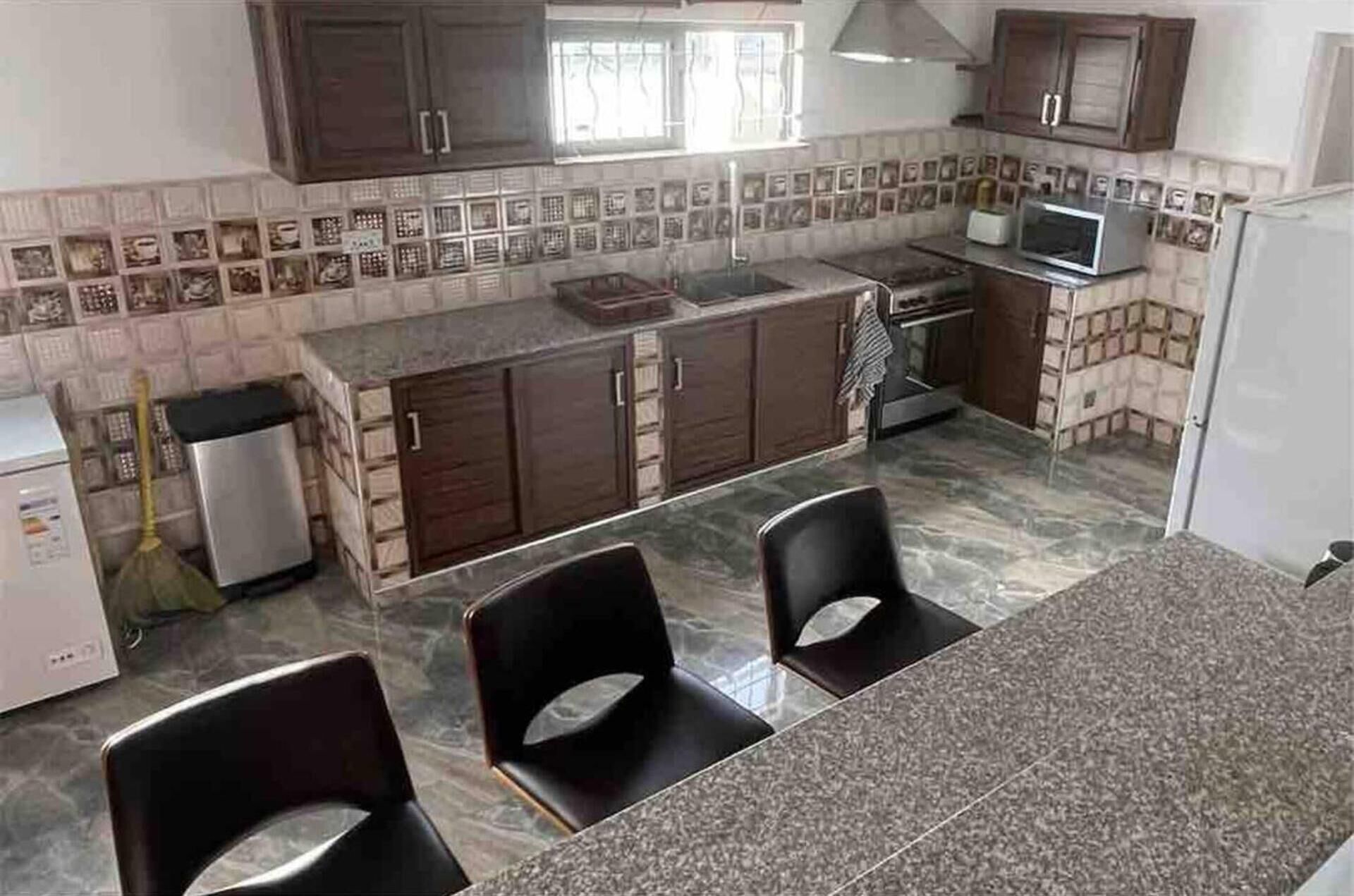 A kitchen with dark wooden cabinets, a tiled backsplash, a microwave, and a sink. There are two black chairs in the foreground.
