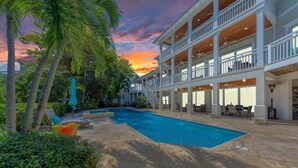 Un área de piscina con un edificio de dos pisos, mobiliario exterior y palmeras.