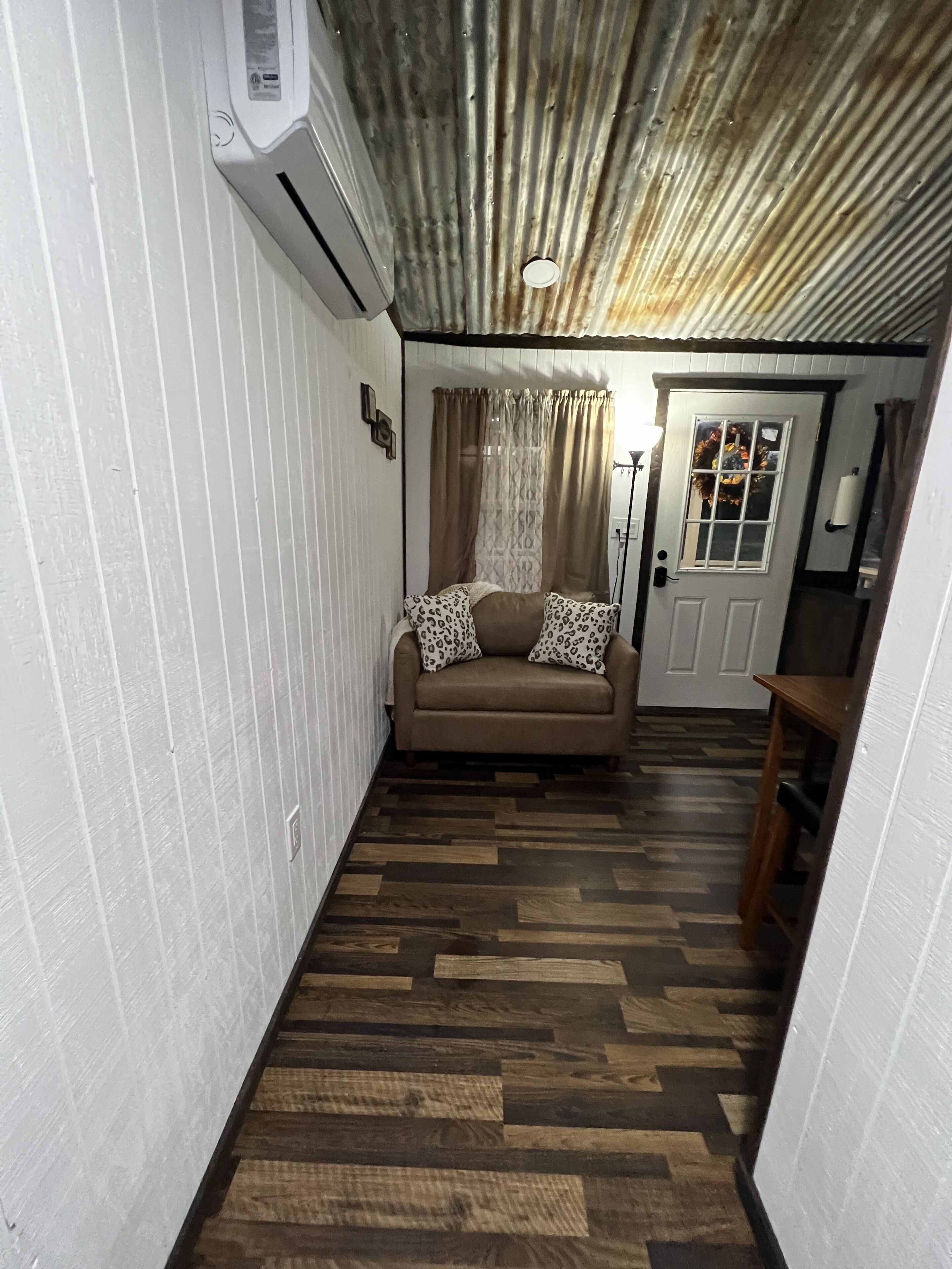 Une pièce avec un plancher en bois, un canapé agrémenté de coussins à motifs, une porte blanche et un plafond orné de plaques de métal ondulées.