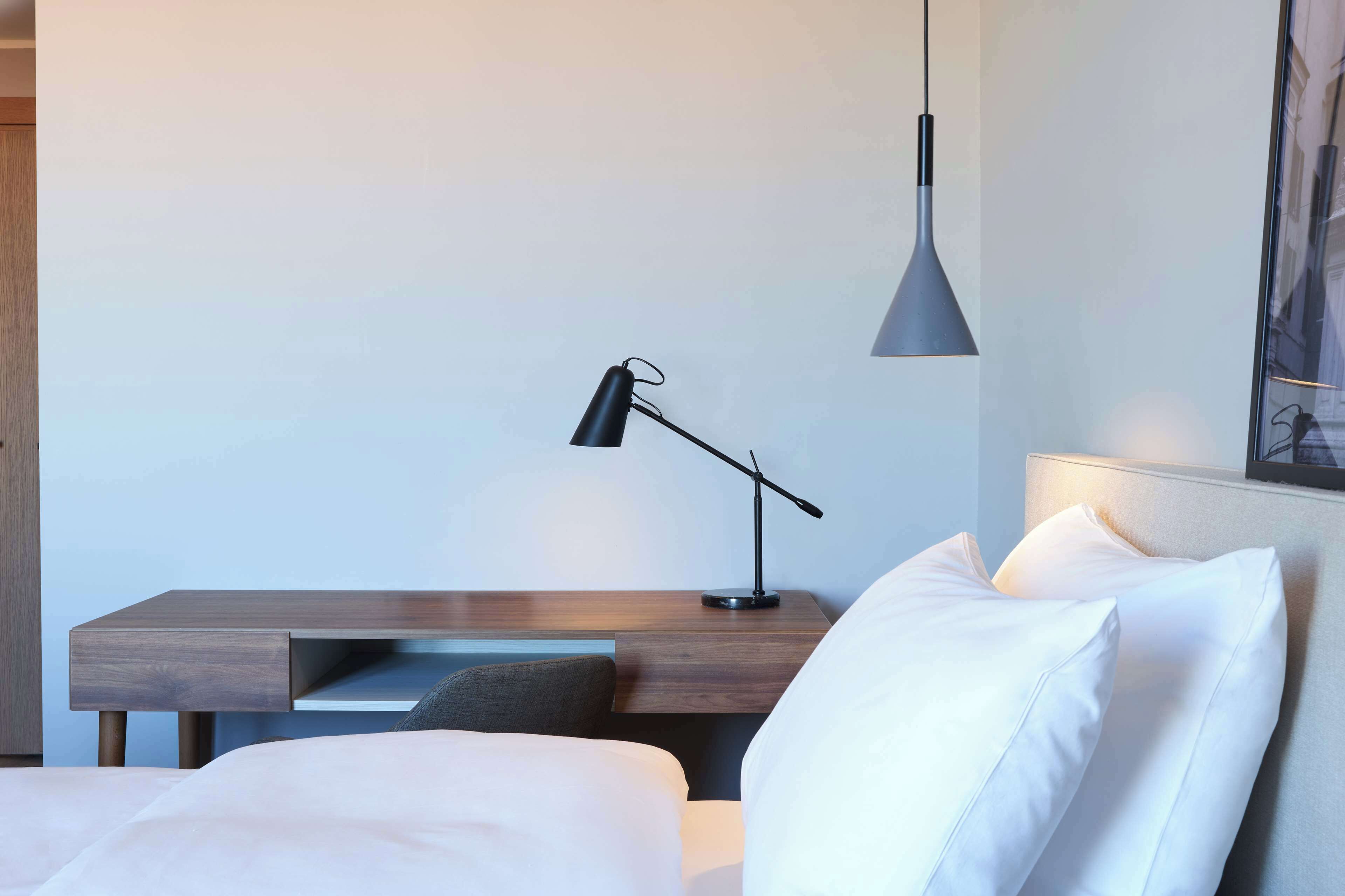 A modern hotel room with a wooden desk, a black desk lamp, a white bed, and a grey pendant light.