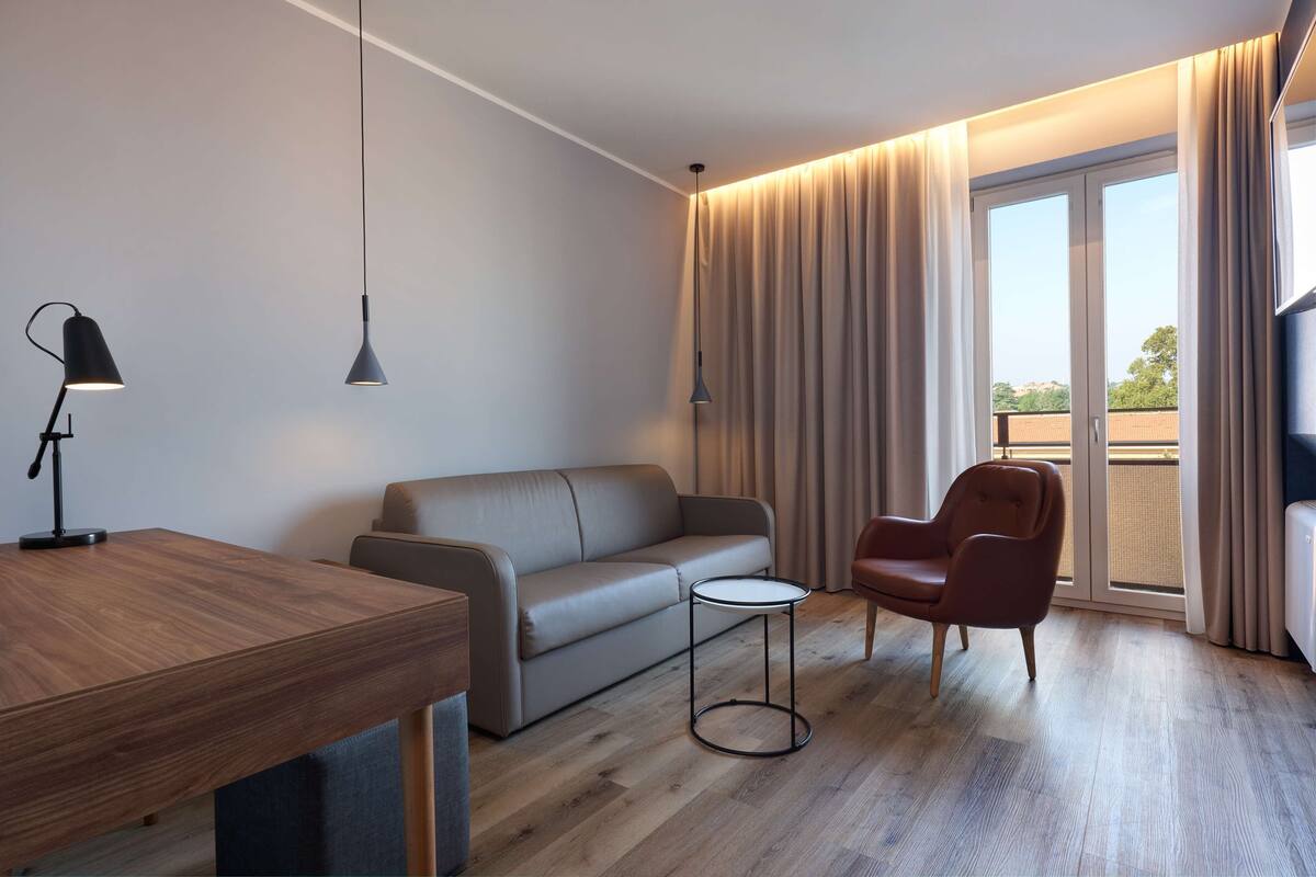 Ein modernes Wohnzimmer mit einem Holztisch, einer Couch, einem kleinen runden Tisch, einem Sessel und einem großen Fenster mit Vorhängen.