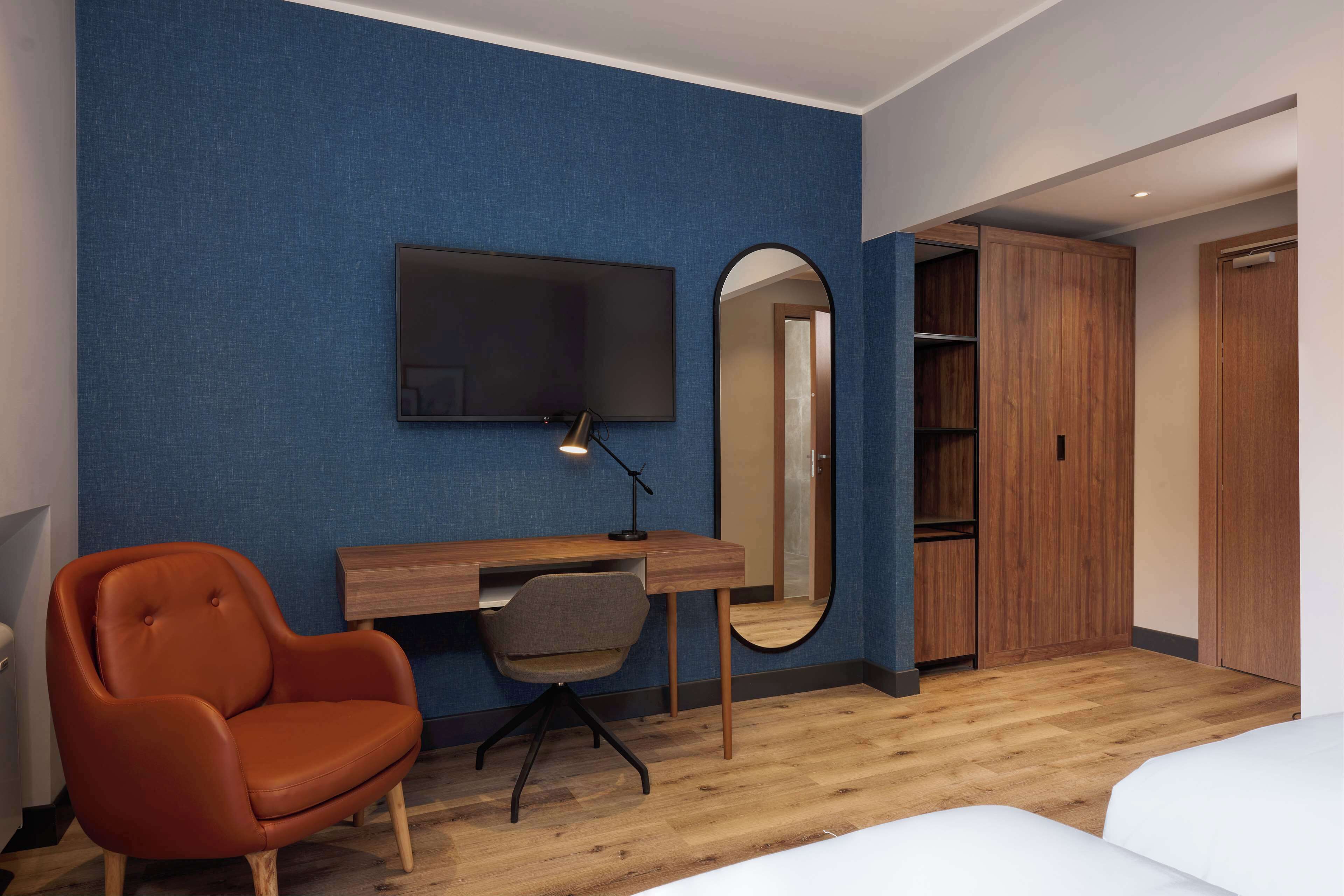 Una camera d'albergo con una scrivania in legno, una televisione, uno specchio, una sedia rossa e un armadio.