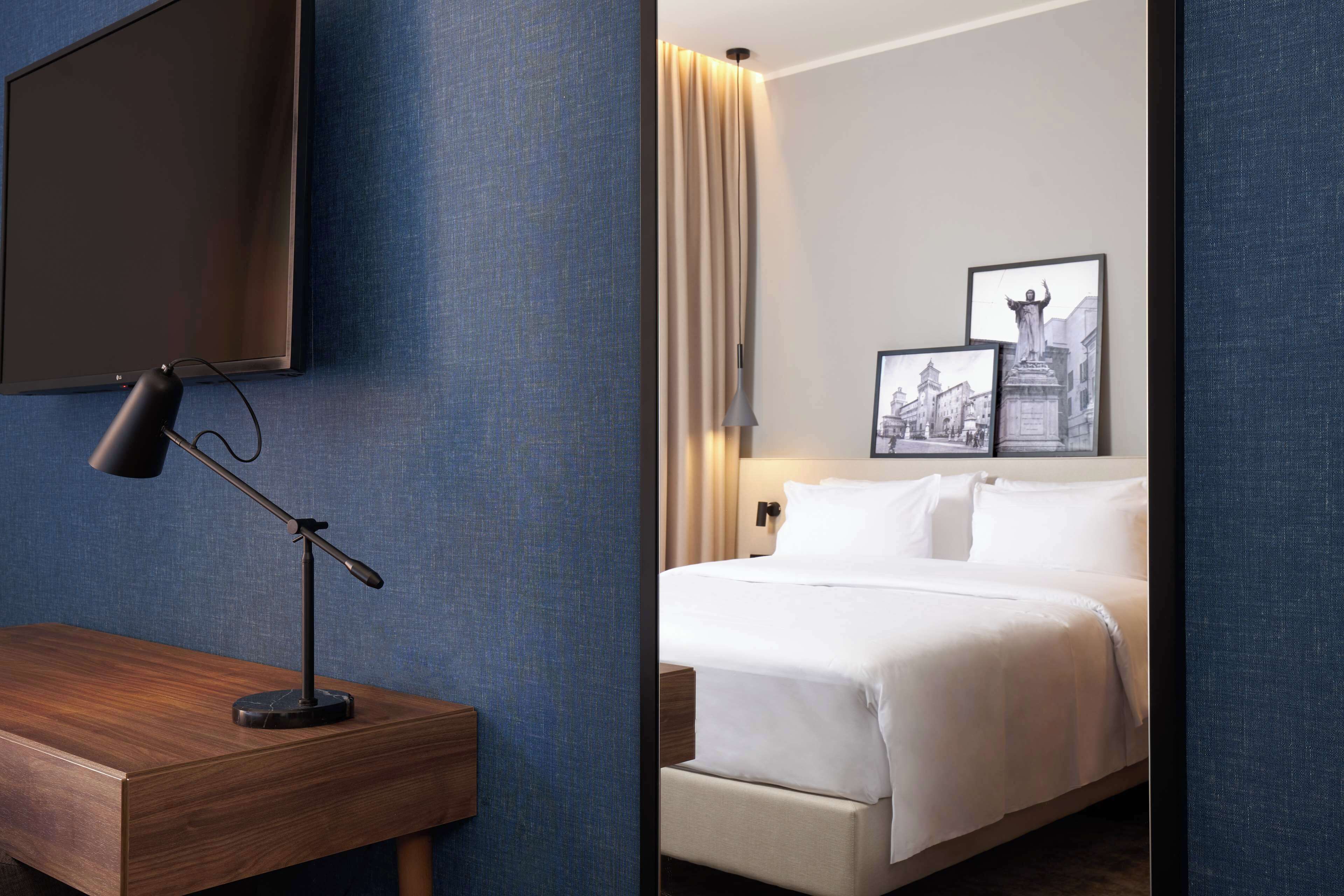 Habitación de hotel con una cama, un escritorio con lámpara y dos cuadros en la pared.