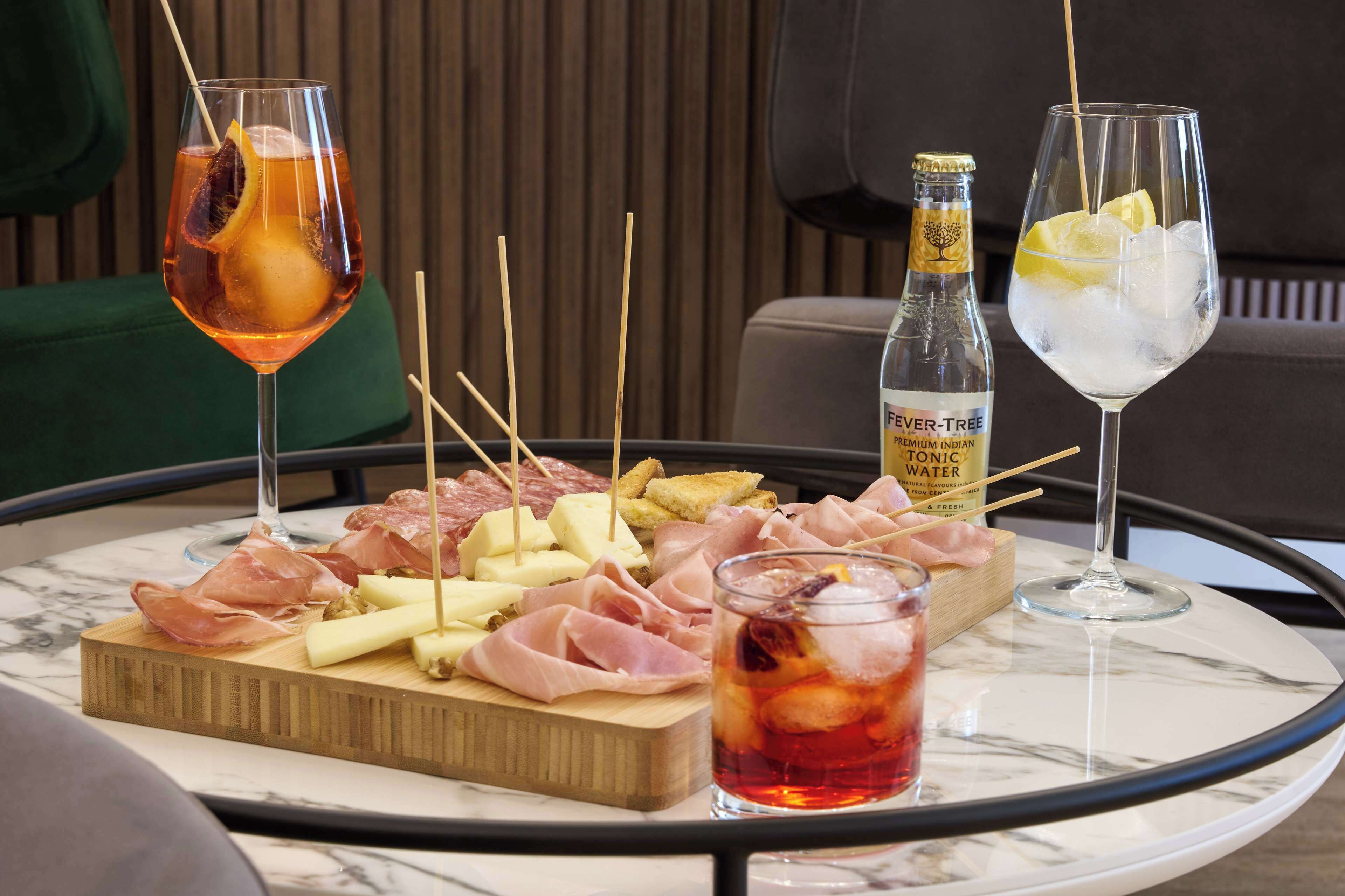 Una mesa con diversos alimentos y bebidas, incluyendo una botella de agua tónica, un vaso de bebida roja y un vaso con rodajas de limón y hielo.