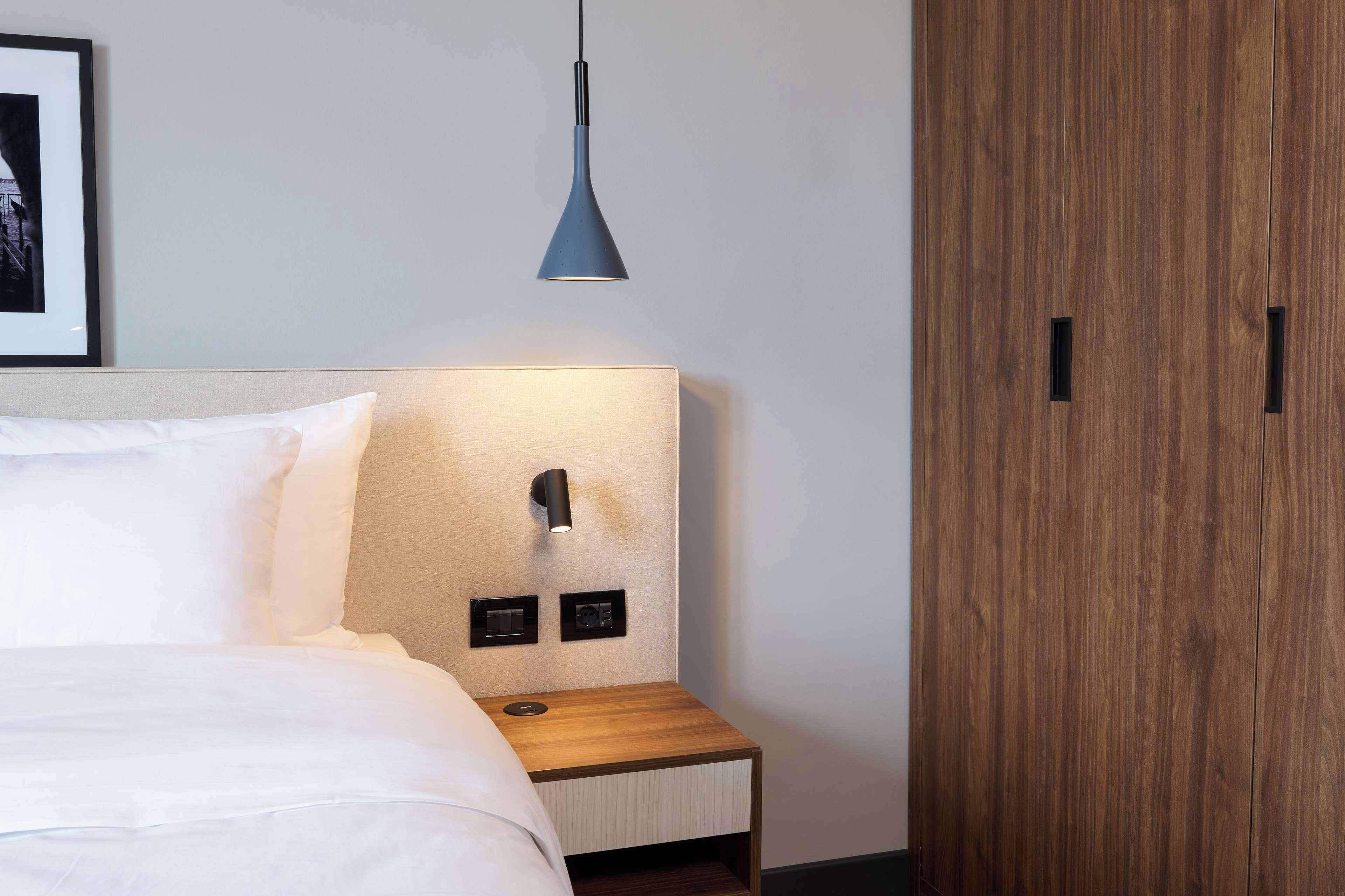 A neatly made bed with white linens, a bedside table with a lamp, and a wooden wardrobe.