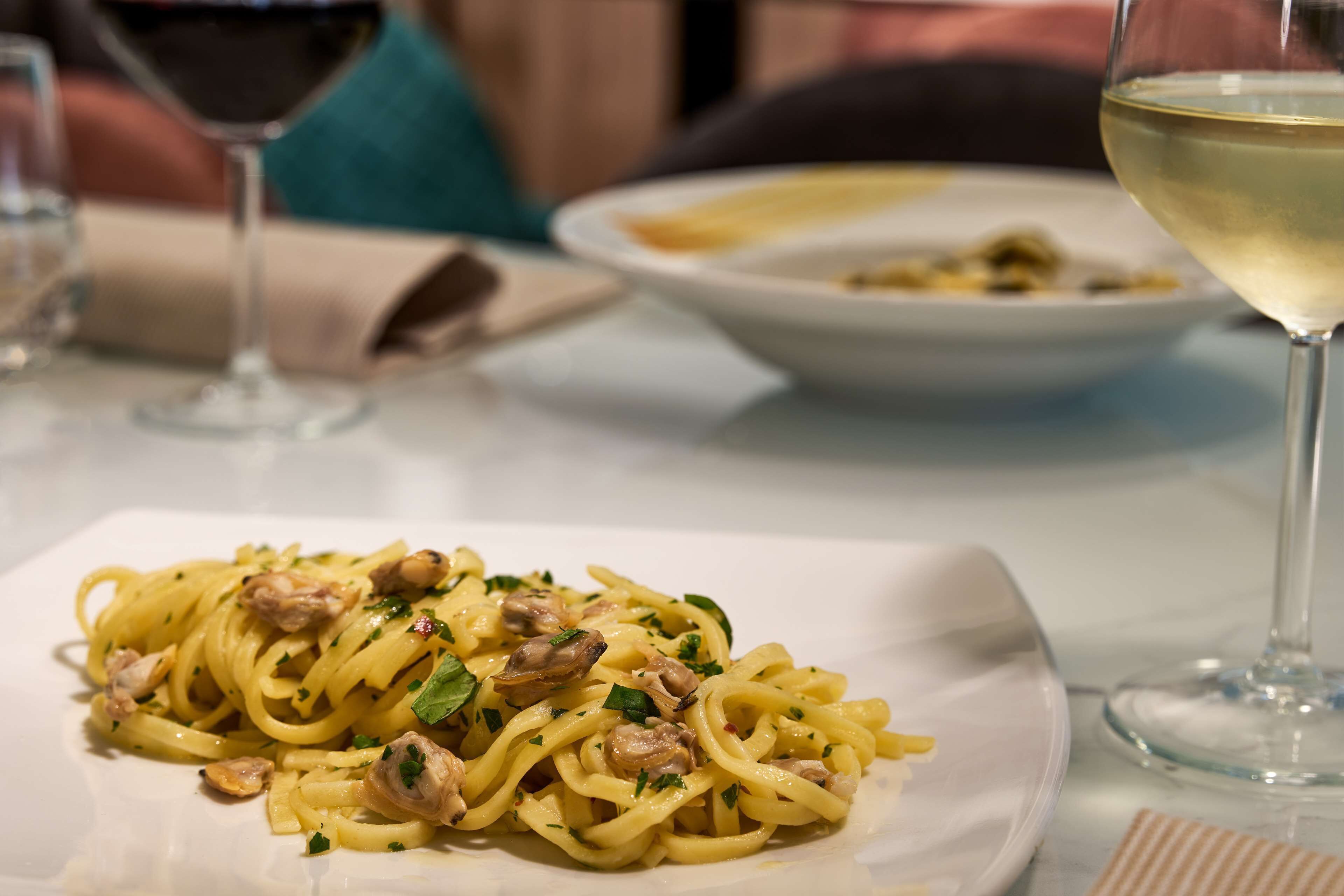 Un piatto di pasta con funghi e erbe aromatiche, un bicchiere di vino bianco e una tovaglietta su un tavolo.