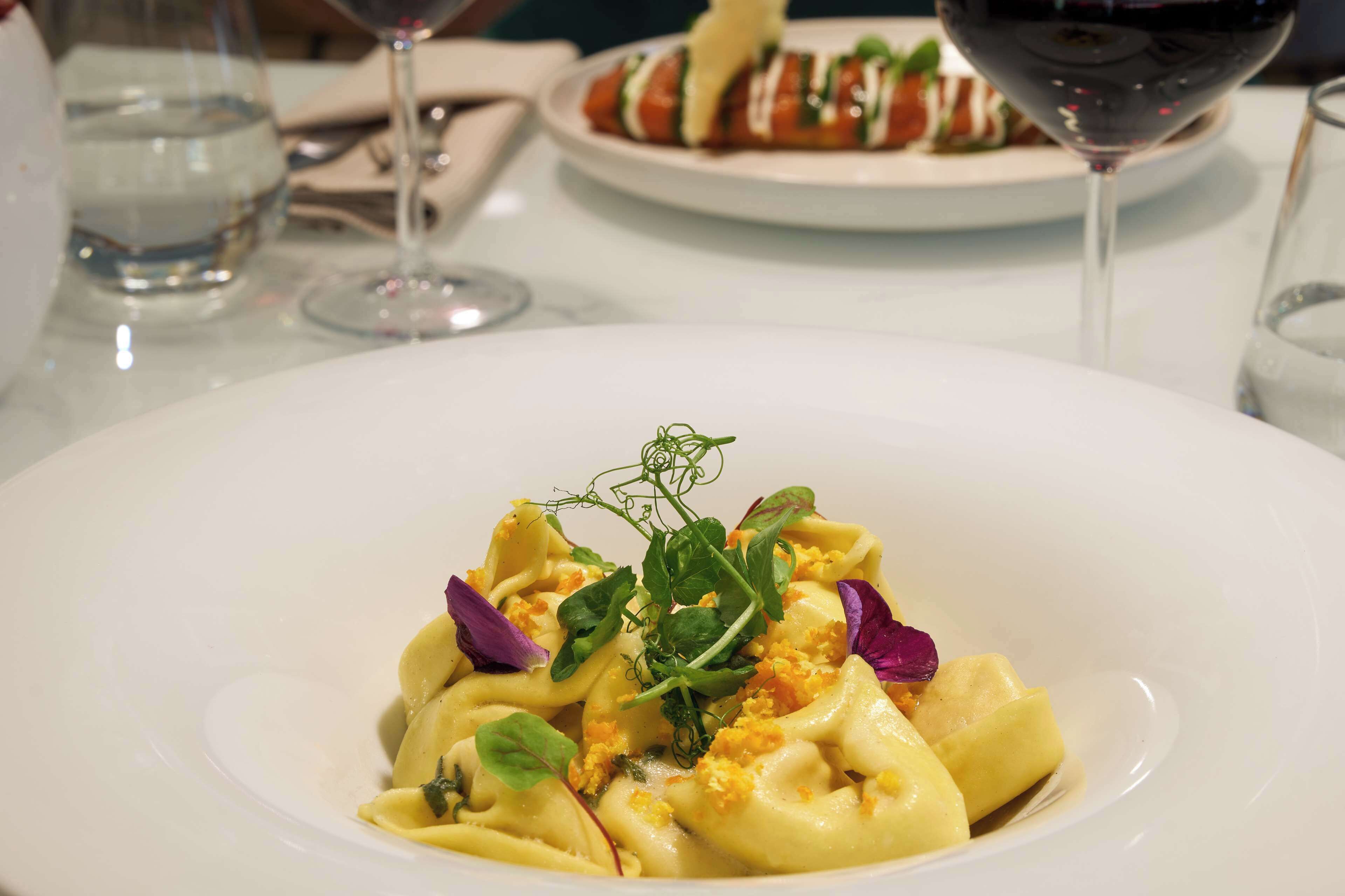 Un plato blanco de pasta con verduras y flores moradas, una copa de vino tinto y un vaso de agua sobre una mesa.
