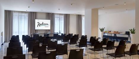 Un salón de conferencias con filas de sillas, una pantalla de proyección y una zona de buffet con comida y bebidas.