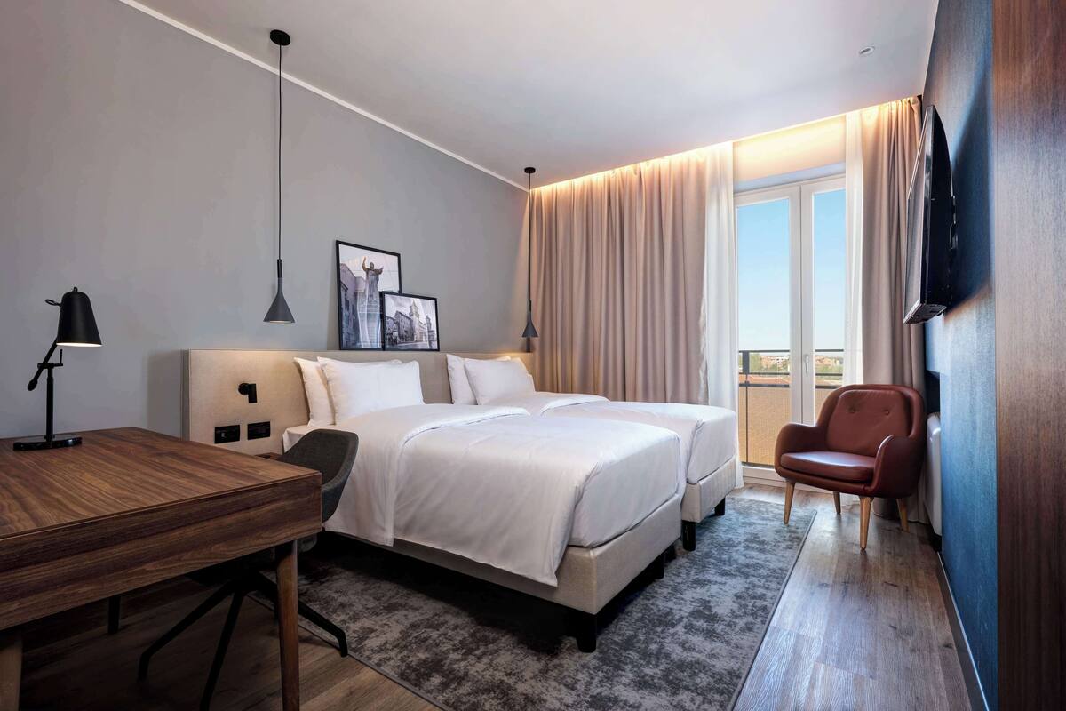 Ein Hotelzimmer mit zwei Betten, einem Schreibtisch, einem Sessel, einem Fernseher und einem großen Fenster mit Vorhängen.
