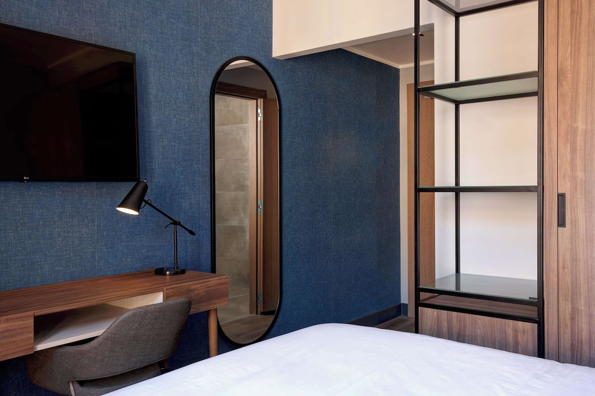 Ein modernes Hotelzimmer mit einem Bett, einem Schreibtisch mit Stuhl, einem Spiegel und einem Kleiderschrank mit Glastüren.