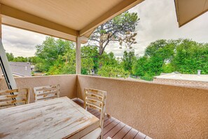 En udendørs terrasse med et bord og stole, en pergola over hovedet og udsigt til træer og et hus i baggrunden.