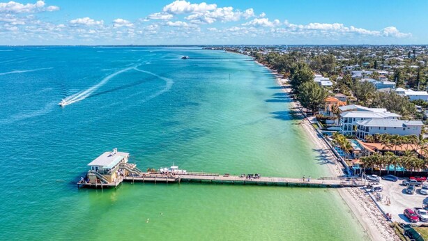 An aerial view of a coastal area with a pier extending into turquoise waters, adjacent to a residential area with houses and a beach. - Anna Maria Island Surfside Bungalow (Holmes Beach)