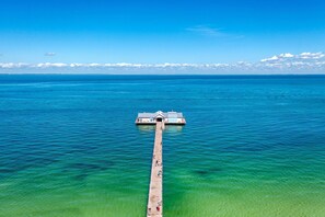 Une vue aérienne d’une jetée menant à un petit quai sur lequel se trouve un bâtiment, le tout étant entouré d’une eau d’un bleu limpide.