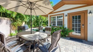 An outdoor patio with a round table and chairs, a large umbrella, and a door with a window. - Anna Maria Island Surfside Bungalow (Holmes Beach)