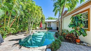 A swimming pool surrounded by lush greenery and a small patio area with a chair and umbrella. - Anna Maria Island Surfside Bungalow (Holmes Beach)
