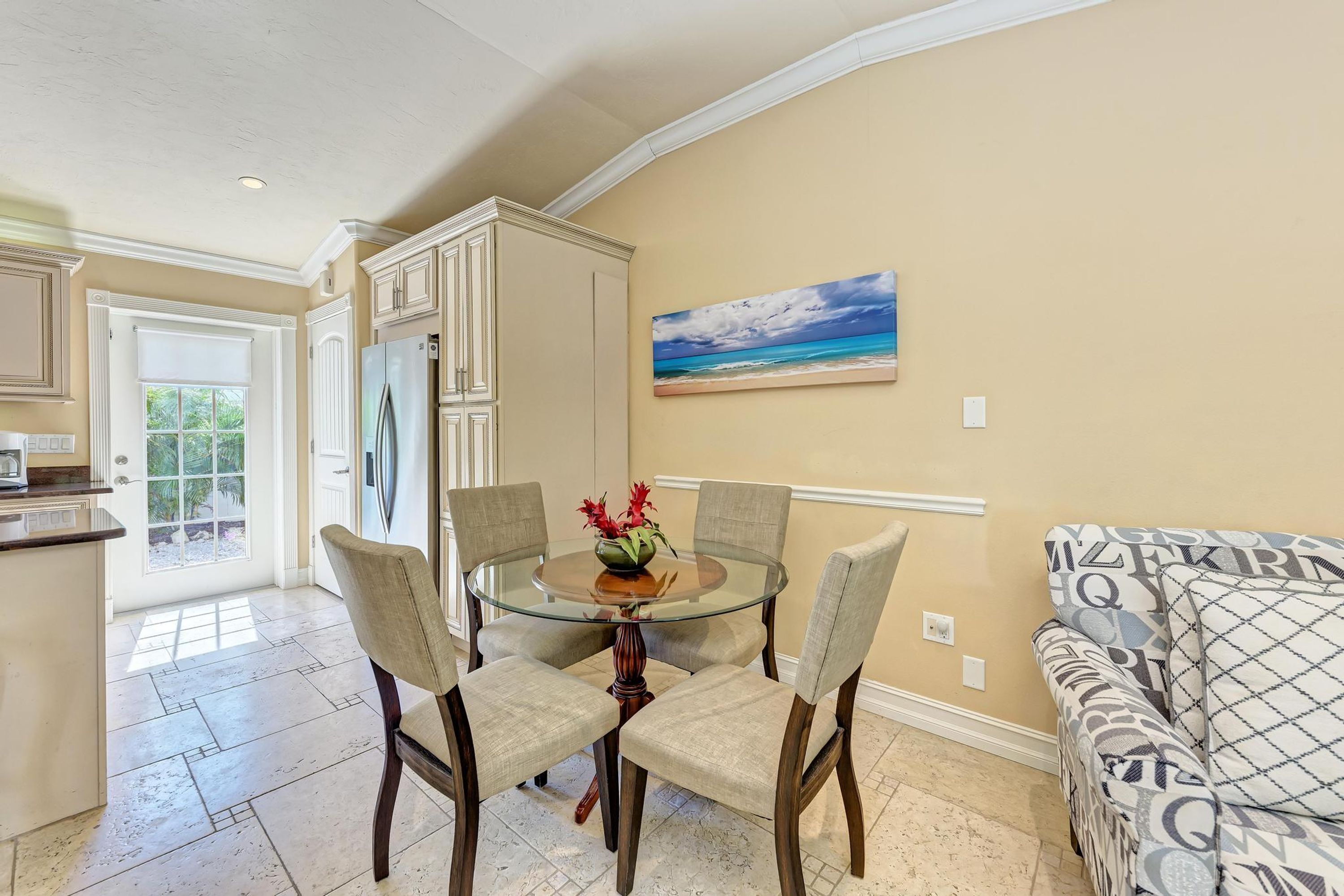 A dining area with a glass-top table surrounded by four chairs, a kitchen with white cabinets, and a painting of a beach scene on the wall.