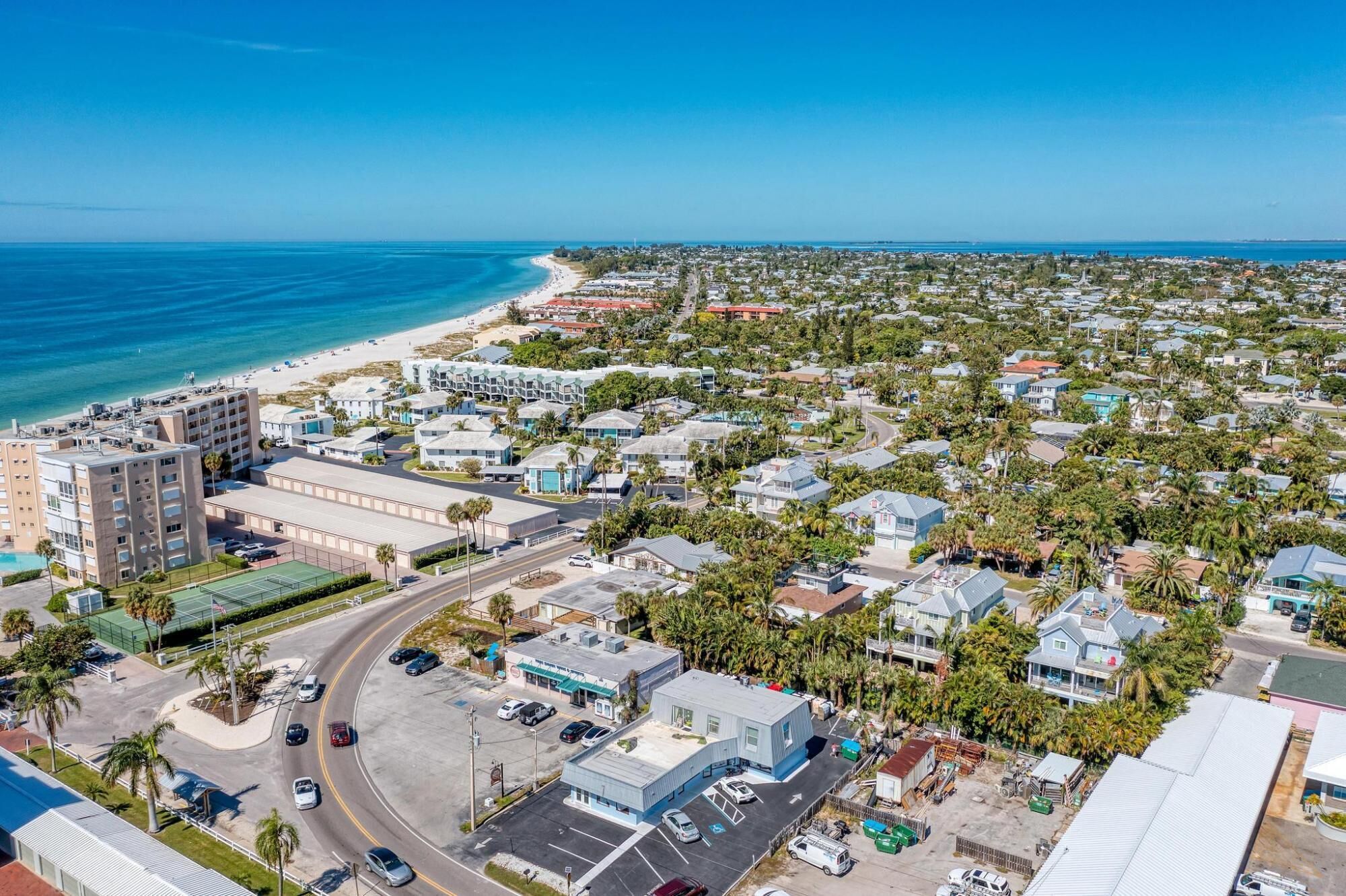 An aerial view of a coastal town with a beach, residential buildings, and a commercial area with parking and a small building with a blue roof.