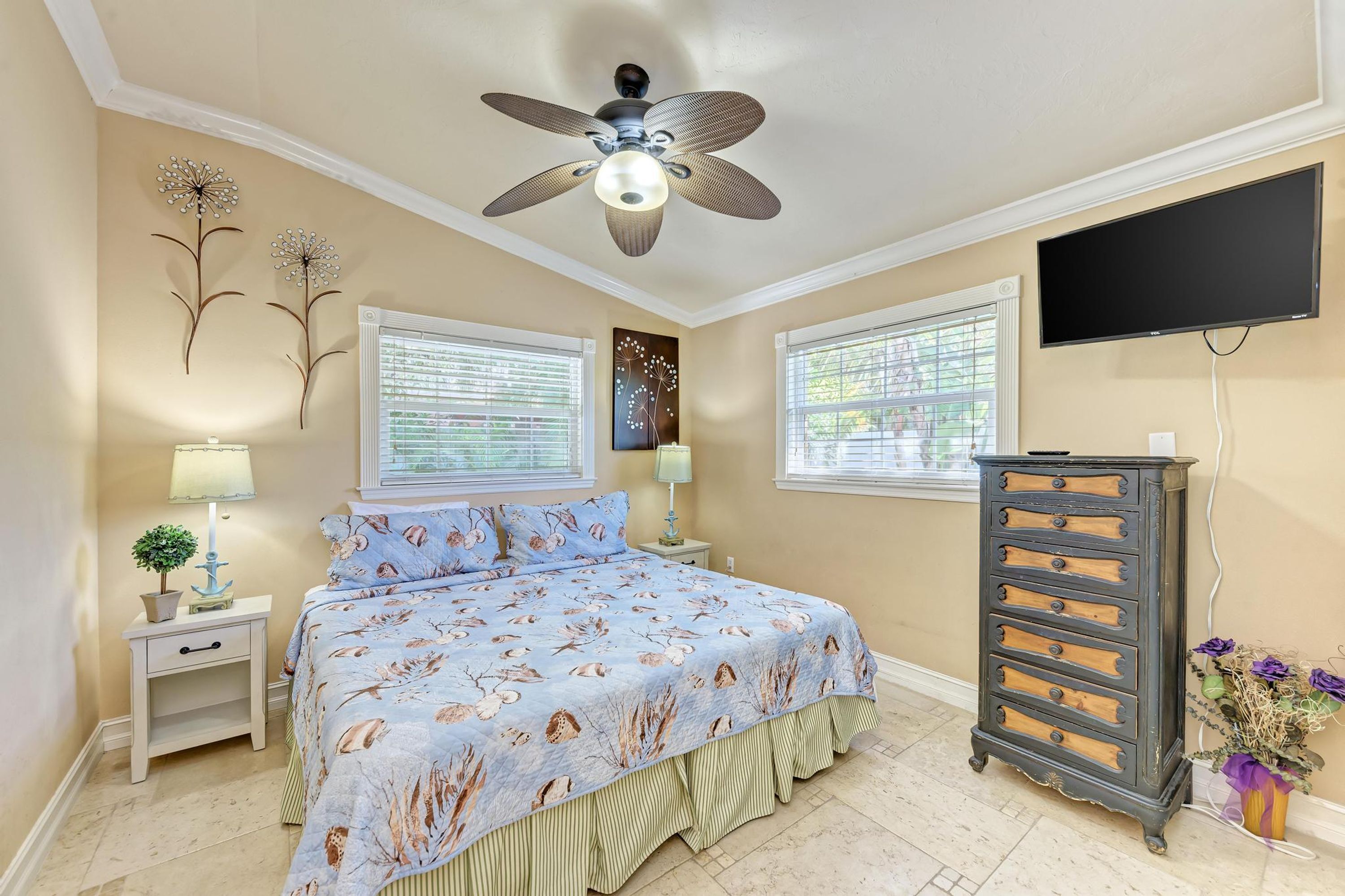 Une chambre à coucher comprenant un lit, deux tables de chevet, un ventilateur de plafond, une commode et un téléviseur fixé au mur.