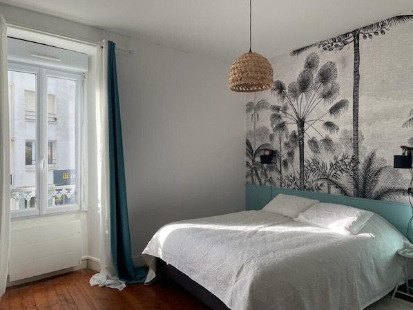 A bedroom with a bed, a window with curtains, a wall mural, and a hanging light fixture.