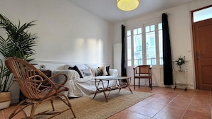 Una habitación con una cama, dos sillones de mimbre, una mesita, una planta en maceta, una ventana con cortinas y una puerta.