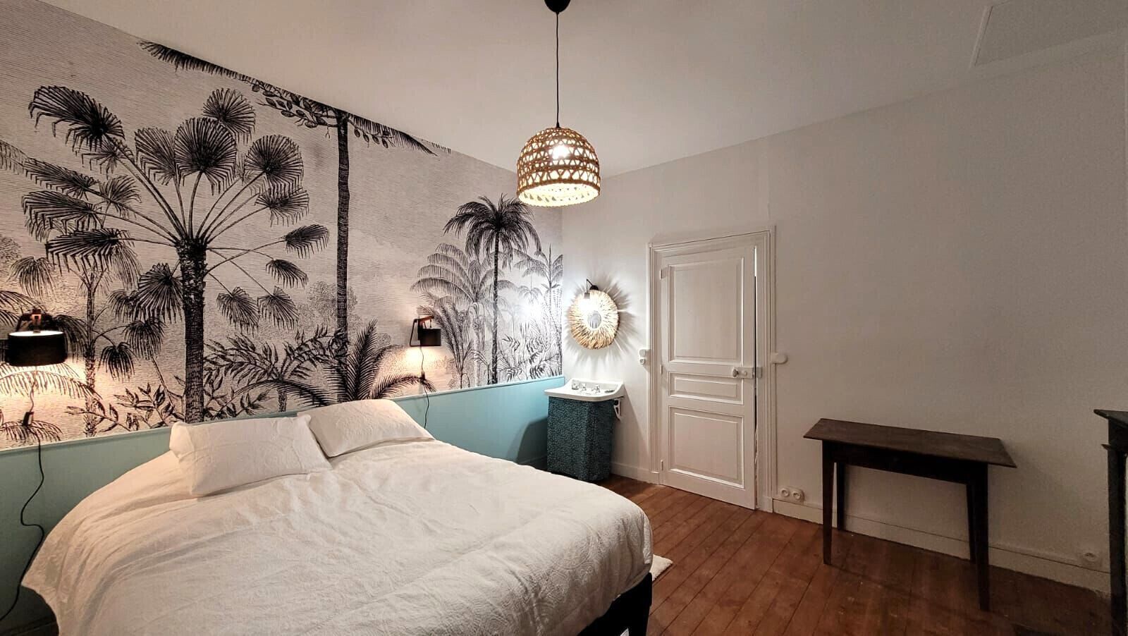 A bedroom with a bed, a wall mural, a hanging light fixture, a bedside table, and a door.