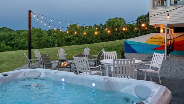 Un bain à remous doté de sièges et illuminé de guirlandes lumineuses, entouré de fauteuils Adirondack, le tout se dressant sur fond d’un bâtiment orné d’une fresque colorée.