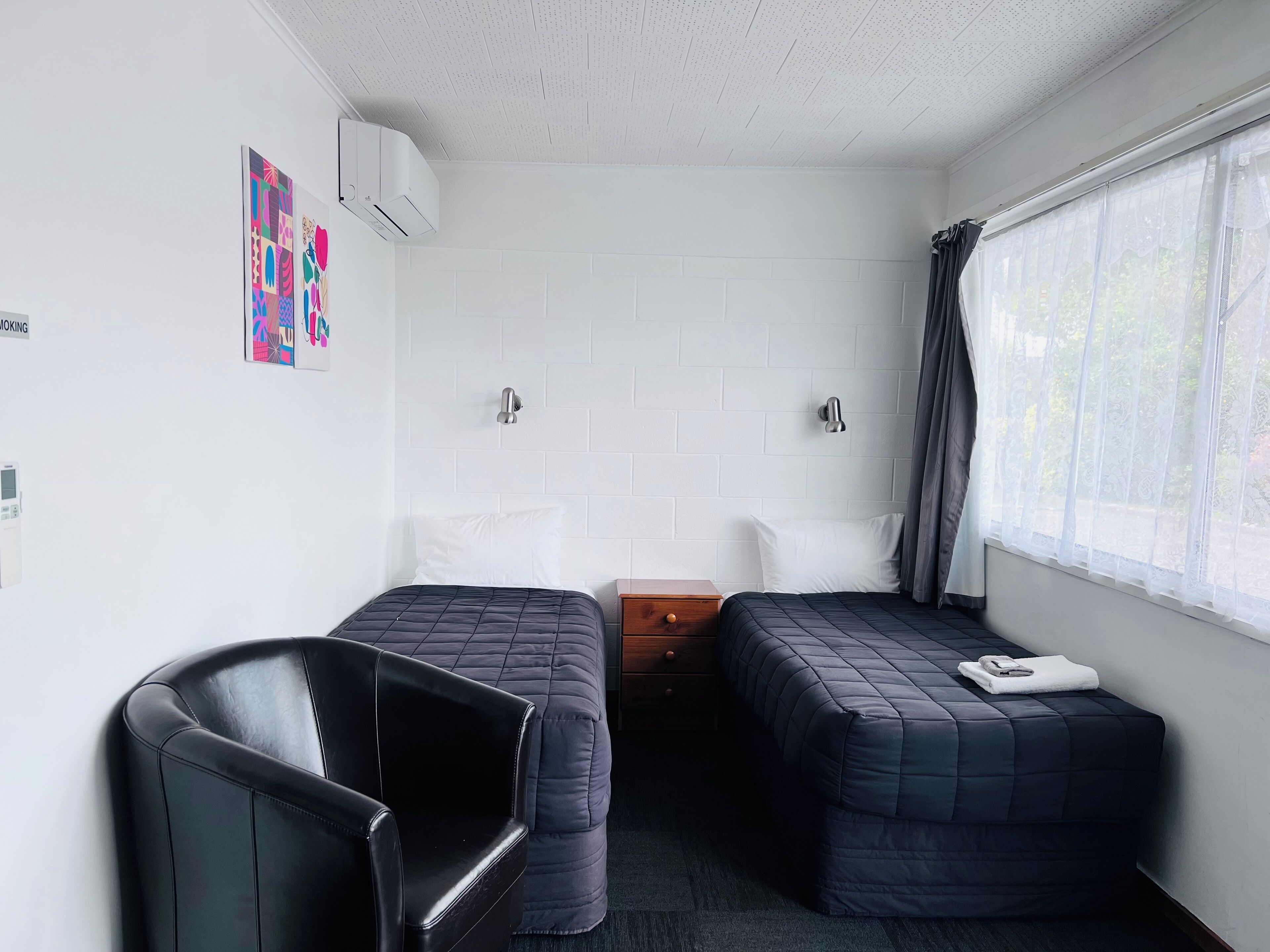 Quarto com duas camas, uma cadeira, uma mesa pequena e uma janela com cortinas.