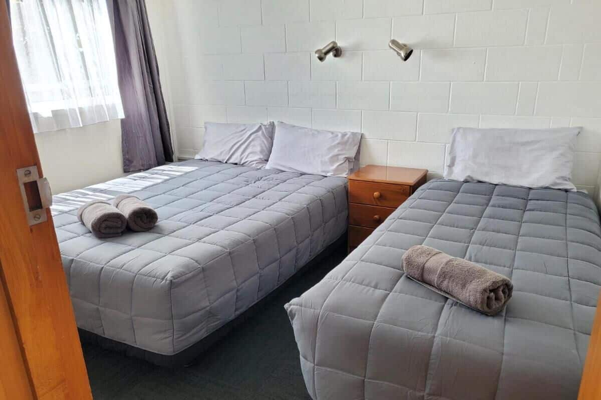 A hotel room with two beds, each with a grey quilted mattress and pillows, a wooden nightstand, and a window with curtains.
