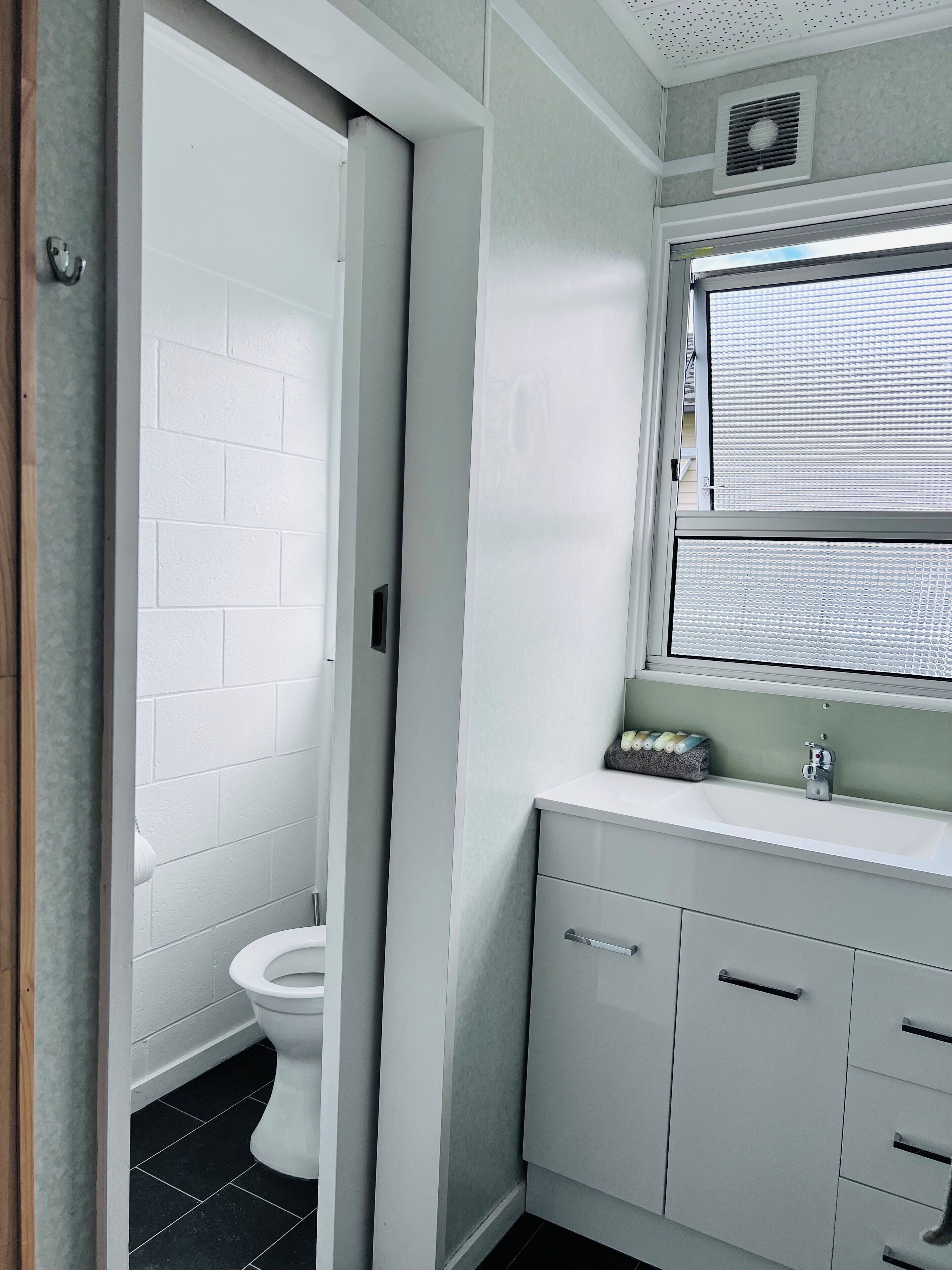 Une salle de bain avec des toilettes blanches, un lavabo placé sous une fenêtre et un meuble avec des tiroirs.