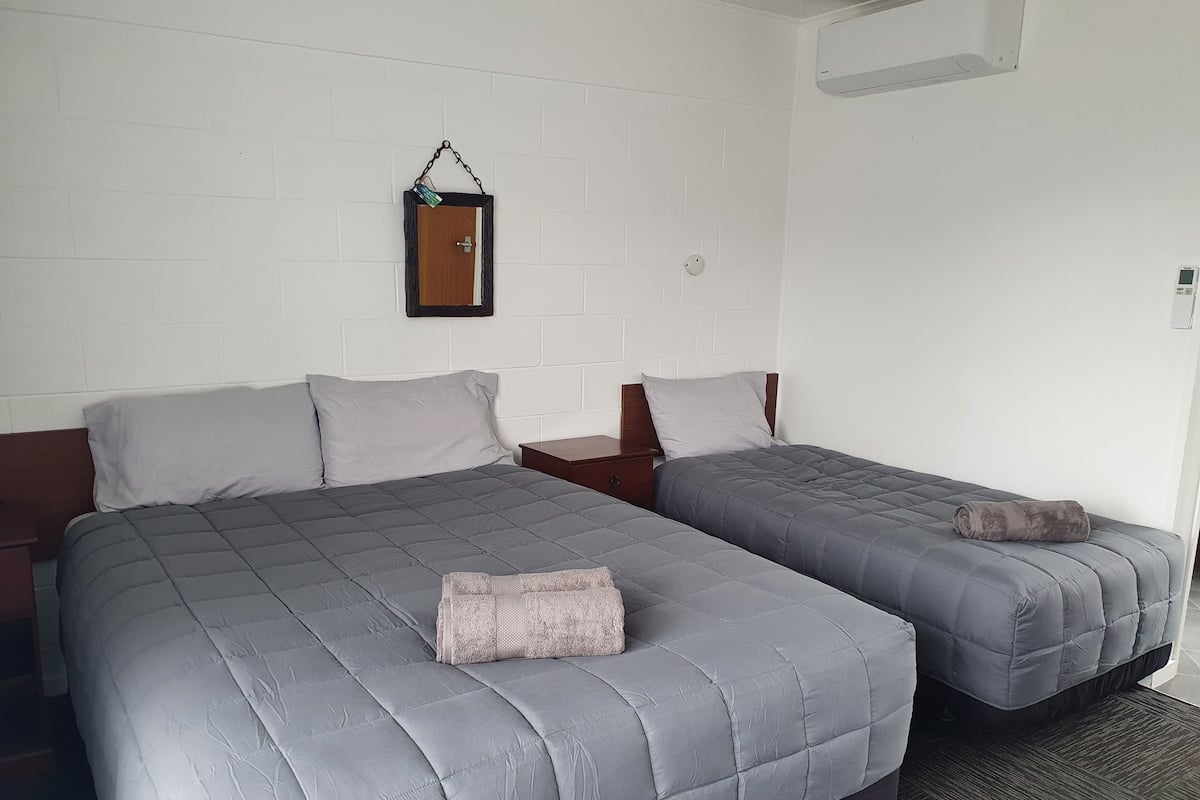 A hotel room with two beds, a small wooden nightstand, a wall-mounted air conditioning unit, and a white brick wall.