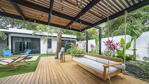 Une terrasse en bois avec une balançoire en bois et un banc, entourée d’une végétation luxuriante et d’un mur blanc.