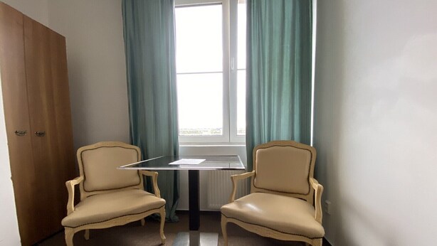 Et værelse med to beige lænestole, et lille bord, et vindue med gardiner og en radiator.