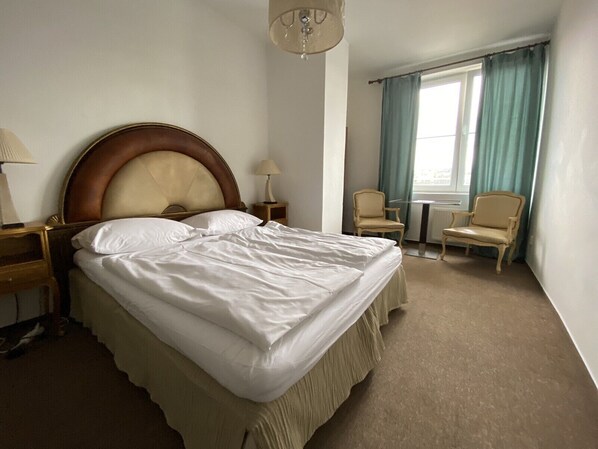 A hotel room with a large bed, two bedside tables, a chair, a small table, and a window with curtains.