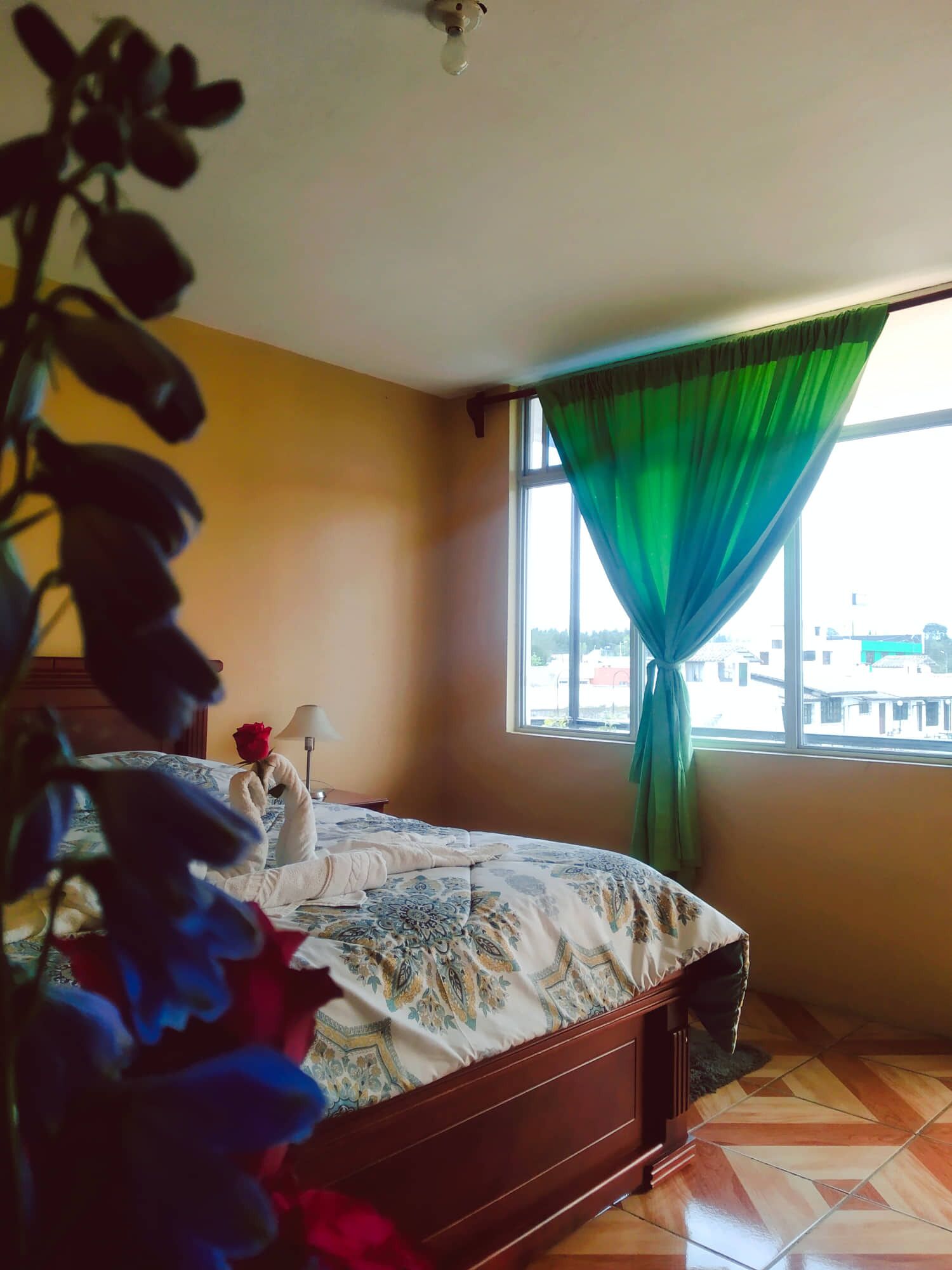 A bedroom with a bed, a window with green curtains, and a view of buildings outside.