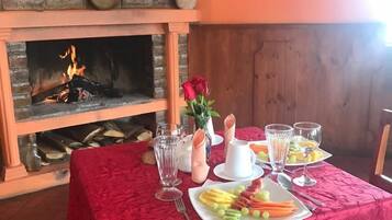 Una mesa de comedor con mantel rojo, platos de fruta y vasos, frente a una chimenea con pared de ladrillo y paredes anaranjadas.
